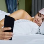Person lying in bed looking at phone, showing how neck pain from phone in bed starts